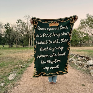 Puede incluir: Una manta de crochet verde con ribetes dorados y una inscripción blanca que dice "Érase una vez, en una tierra que hace mucho tiempo se convirtió en ceniza, vivía una joven princesa que amaba mucho su reino."