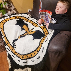 K&ouml;nnte beinhalten: Ein kleiner Junge sitzt in einem braunen Ledersessel und liest ein "World of Reading" Buch &uuml;ber Star Wars. Er tr&auml;gt ein schwarz-graues Shirt mit dem Wort "adidas" darauf. Er sitzt auf einer schwarz-wei&szlig;en Decke mit einem gelben Muster.