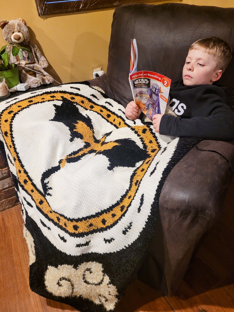 K&ouml;nnte beinhalten: Ein kleiner Junge sitzt in einem braunen Ledersessel und liest ein "World of Reading" Buch &uuml;ber Star Wars. Er tr&auml;gt ein schwarz-graues Shirt mit dem Wort "adidas" darauf. Er sitzt auf einer schwarz-wei&szlig;en Decke mit einem gelben Muster.