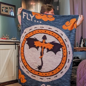 K&ouml;nnte beinhalten: Eine blaue geh&auml;kelte Decke mit einem orangenen Kreis, der eine schwarze Drachen-Silhouette und den Text "FLY...OR DIE" zeigt.