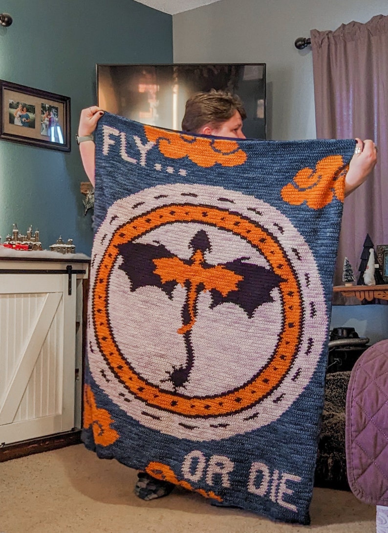 K&ouml;nnte beinhalten: Eine blaue geh&auml;kelte Decke mit einem orangenen Kreis, der eine schwarze Drachen-Silhouette und den Text "FLY...OR DIE" zeigt.