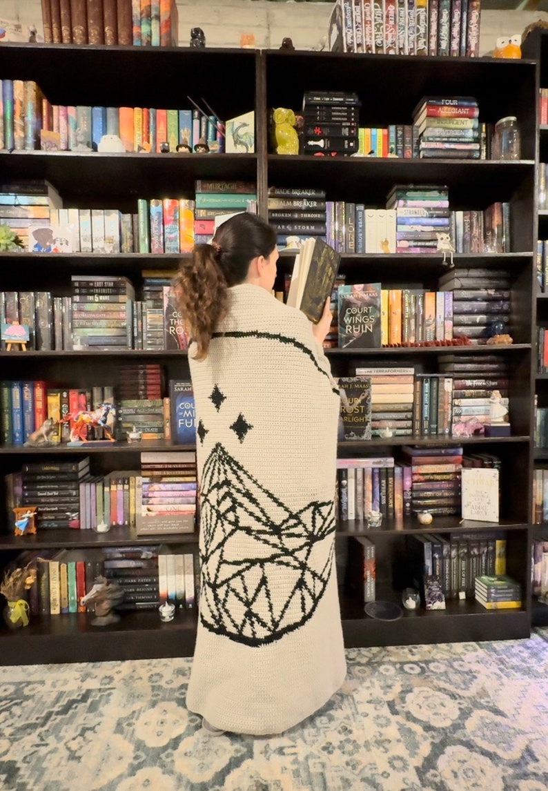 May include: A person standing in front of a bookshelf filled with books, wearing a cream-colored crocheted blanket with a black geometric mountain design and stars.