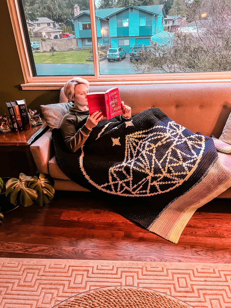 May include: A person is sitting on a couch reading a book titled "A Court of Wings and Ruin". They are wrapped in a black and white geometric patterned blanket. The couch is beige with button tufting. The person is wearing a green shirt.