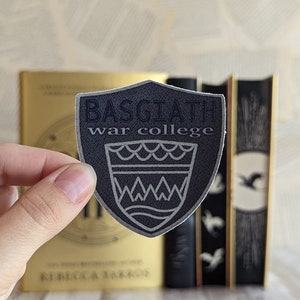 Pode incluir: Um autocolante em forma de escudo com o texto "BASGIATH war college" numa textura cinzenta escura semelhante a ganga. O autocolante apresenta um emblema branco de uma coroa e ondas. O autocolante é segurado em frente a um livro dourado e dois livros pretos.