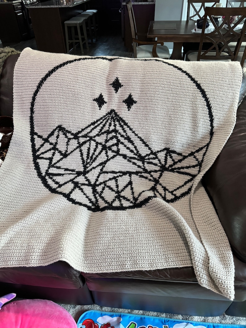 May include: A crocheted throw blanket with a black and white geometric mountain design and three stars in a circle.