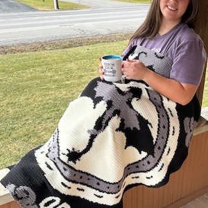 K&ouml;nnte beinhalten: Eine Frau sitzt auf einer Veranda mit einer geh&auml;kelten Decke mit einem schwarz-wei&szlig;en Design mit dem Text "OR DIE". Sie h&auml;lt eine wei&szlig;e Tasse mit blauem Rand, auf der "Crocheting keeps me from unraveling" steht.
