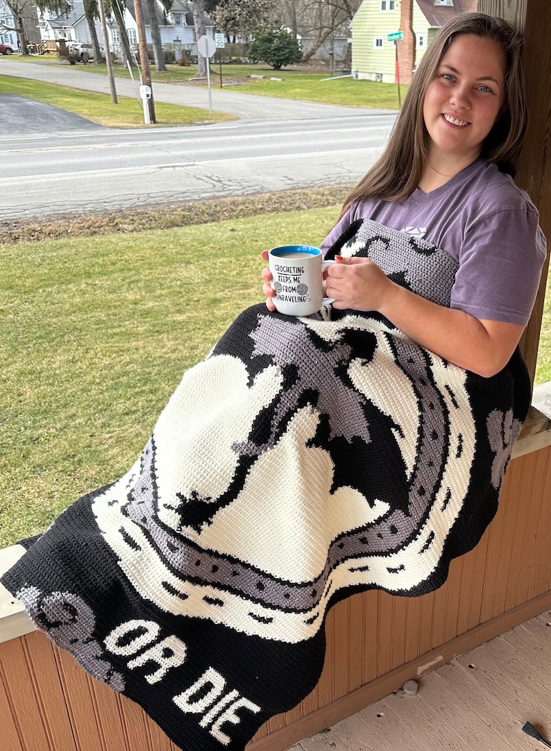 K&ouml;nnte beinhalten: Eine Frau sitzt auf einer Veranda mit einer geh&auml;kelten Decke mit einem schwarz-wei&szlig;en Design mit dem Text "OR DIE". Sie h&auml;lt eine wei&szlig;e Tasse mit blauem Rand, auf der "Crocheting keeps me from unraveling" steht.