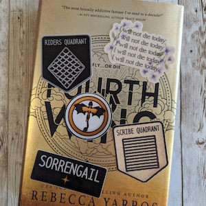Op de afbeelding: Een goudkleurig boek met de titel "Fourth Wing" van Rebecca Yarros, versierd met verschillende stickers. De stickers bevatten zinnen als "I will not die today" en "Riders Quadrant" en "Sorrengail". Het boek heeft een citaat van Tracy Wolff.
