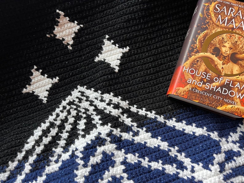 May include: A black and white crochet blanket with a blue and white pattern at the bottom. The blanket features a mountain design and three stars. A book titled "House of Flame and Shadow" by Sarah J. Maas is on top of the blanket.