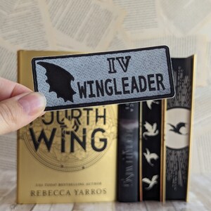 Może przedstawiać: Prostokątna naszywka z napisem "IV WINGLEADER" i czarnym motywem skrzydła nietoperza. Naszywka jest trzymana przed złotą książką zatytułowaną "Fourth Wing" autorstwa Rebeccy Yarros i innymi książkami z czarnymi grzbietami.