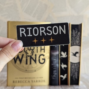 Puede incluir: Una pegatina rectangular negra con el nombre "RIORSON" en letras blancas y tres estrellas doradas. La pegatina se sostiene frente a un libro dorado con el título "FOURTH WING" y otros libros con lomos negros.