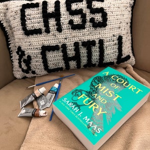 May include: A white and black crocheted pillow with the text "CH55 & CHILL" on a brown couch. A teal book with a silver and black illustration of a bird on the cover is on the couch. The book title is "A Court of Mist and Fury" by Sarah J. Maas.