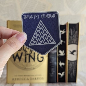 Puede incluir: Una pegatina azul marino y blanca de Infantry Quadrant sostenida frente a una pila de libros. La pegatina tiene un diseño geométrico y el texto "Infantry Quadrant". Los libros tienen cubiertas doradas y negras.