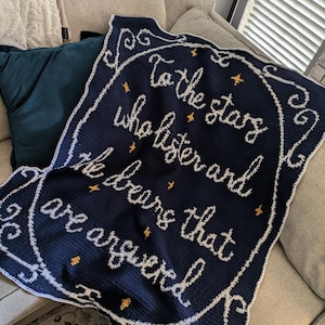 Puede incluir: Una manta de crochet azul marino con puntadas blancas y estrellas doradas. La manta presenta el texto "To the stars who listen and the bears that are answered."
