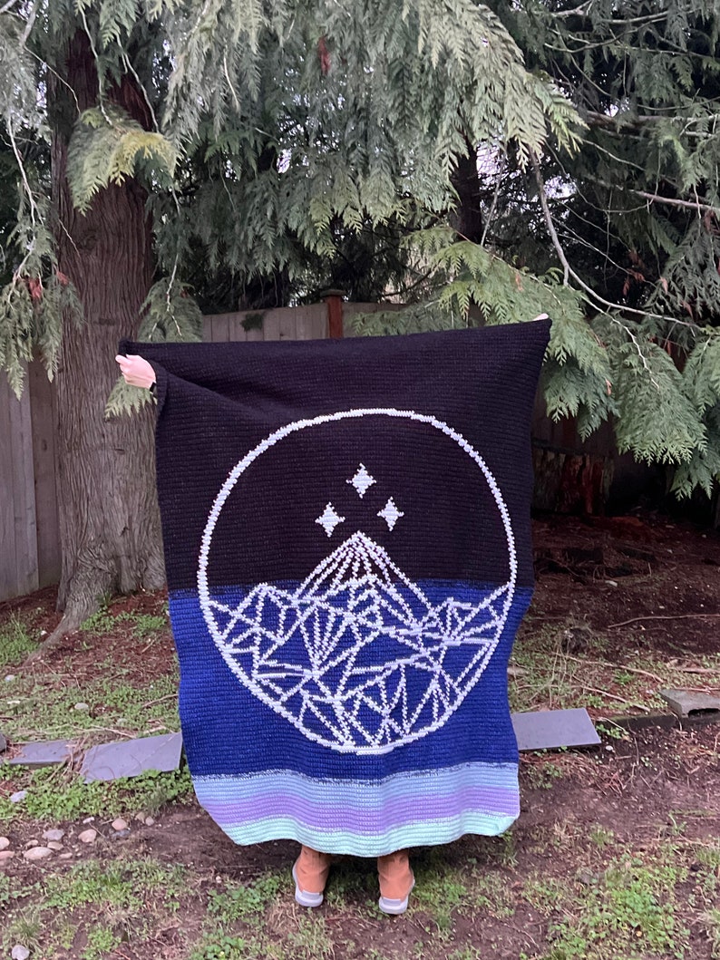 May include: A black, blue, and purple crocheted blanket with a white mountain and star design. The blanket is folded in half and held by a person.