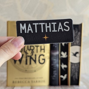 Peut inclure: Un écusson rectangulaire noir avec le nom "MATTHIAS" en lettres blanches et une étoile dorée. L'écusson est tenu devant une pile de livres, dont un avec le titre "FOURTH WING".