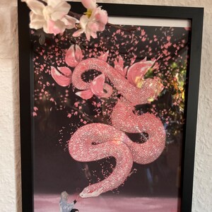 May include: Framed art print depicting a pink snake with a floral design, set against a dark background with falling pink petals. A figure sits below the snake. The print is in a black frame, with cherry blossom branches above.