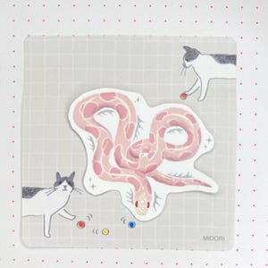 Pode incluir: Um adesivo com uma cobra rosa com um padrão branco sobre um fundo cinza claro com dois gatos brancos e pontos coloridos. O adesivo diz "MIDORI" no canto inferior direito.