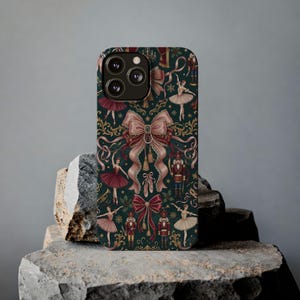 Puede incluir: Una funda de teléfono con un fondo verde oscuro que presenta un patrón repetido de bailarinas de ballet, lazos, cascanueces y elementos decorativos en tonos de rojo, rosa y dorado. La funda tiene un acabado brillante y un recorte para triple lente.