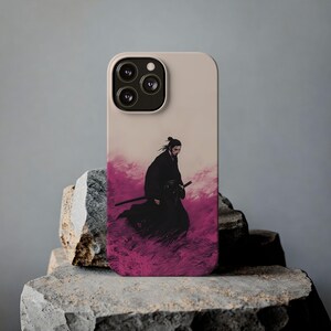 May include: A beige phone case featuring a samurai warrior illustration. The artwork depicts a figure in black robes with a sword, set against a backdrop of pink and purple hues. The phone case has a camera module with three lenses.