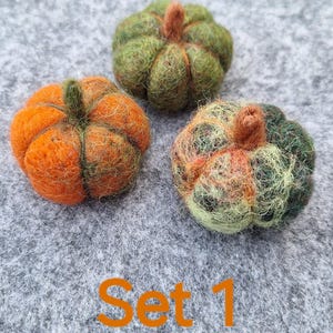 Könnte beinhalten: Drei handgefertigte Filzkürbisse in Orange- und Grüntönen mit braunen Stielen, angeordnet auf einer grauen Filzoberfläche. Der Text "Set 1" ist orangefarben dargestellt. Diese dekorativen Kürbisse sind ideal für die Herbstdekoration.