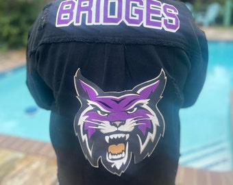 May include: Black denim jacket with a purple and white "BRIDGES" patch on the back and a purple and white wildcat mascot patch on the front.