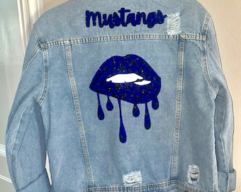 May include: A light blue denim jacket with distressed details. The back of the jacket features a blue glitter lips design with a leopard print pattern and the word "Mustangs" in blue glitter.