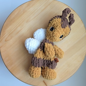 May include: A handmade plush bee toy with brown and tan stripes, white wings, and black eyes. The bee is made of soft, textured yarn and has small antennae. A small brown tag is attached to the bee's body. The toy is lying on a wooden surface.