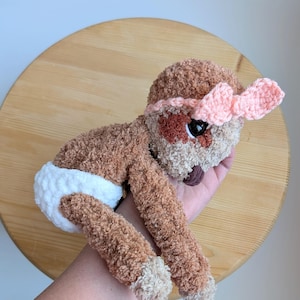 May include: A handmade crochet baby doll with brown fur, a pink bow, and a white diaper. The doll has a brown and tan face with black eyes and a small, dark brown nose. The doll is lying on its stomach.