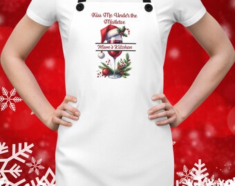 Puede incluir: Delantal blanco con correas negras y un diseño festivo. El delantal presenta el texto "Kiss Me Under the Mistletoe" encima de una copa de vino con un gorro de Papá Noel y las palabras "Mom's Kitchen" debajo.