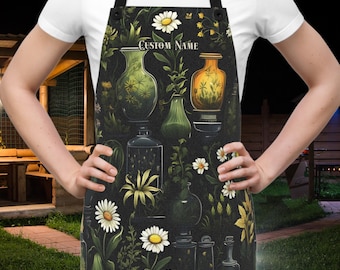 May include: Black apron featuring a botanical design with daisies, vases, and foliage. The apron has a black neck strap and waist ties. The text "CUSTOM NAME" is printed on the apron. The apron is designed for kitchen or craft use.