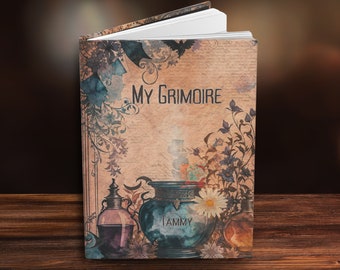 May include: A closed book with a vintage design. The cover features a blue cauldron with the word "TAMMY" on it, surrounded by flowers and herbs. The title "MY GRIMOIRE" is written in black lettering on the cover.