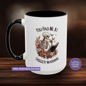 Può includere: Una tazza da caffè in ceramica bianca con bordo e manico neri. La tazza presenta un'illustrazione in bianco e nero di uno scheletro che legge un libro con farfalle che volano intorno a lui. Il testo "You had me at trigger warning" è stampato sulla tazza.