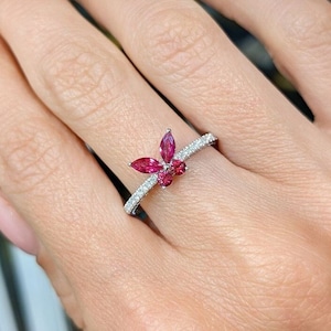 Op de afbeelding: Een zilveren ring met een cluster van rode edelstenen in de vorm van een vlinder. De ring is bezet met kleine diamanten rond de band.