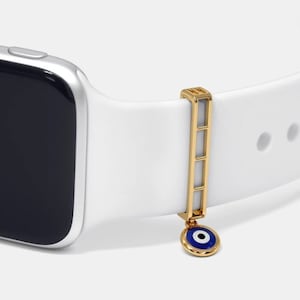May include: A gold-coloured watch band charm with a blue and white evil eye pendant. The charm is attached to a white watch band.