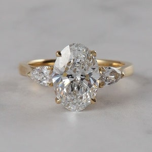 Puede incluir: Un anillo de compromiso de oro con un gran diamante central de talla ovalada y dos piedras laterales en forma de pera. El anillo está engastado en una sencilla banda de oro, mostrando el brillo de los diamantes.