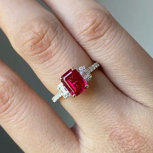 Könnte beinhalten: Ein goldener Ring mit einem großen, rechteckigen, leuchtend roten Edelstein als Mittelpunkt. Der Ring ist mit kleineren, klaren Edelsteinen auf beiden Seiten des Hauptsteins verziert, die in einem filigranen Band gefasst sind.