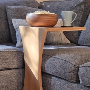 May include: A wooden C-table with a curved base and a small tabletop. A wooden bowl filled with popcorn and a light blue mug sit on the table. The table is next to a gray couch.