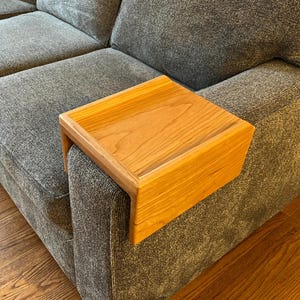 May include: A wooden armrest tray made of light brown wood with a smooth finish. The tray is attached to a gray couch and is perfect for holding a drink or snack.