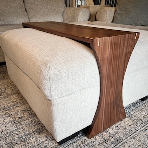 May include: A modern, walnut-toned wooden ottoman tray table. The tray rests on a light-colored upholstered ottoman. The table has a curved, architectural design and is likely used for placing drinks or snacks.