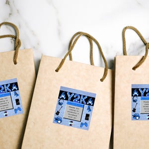 Puede incluir: Tres bolsas de regalo de papel color beige con asas de cuerda marrón. Cada bolsa tiene una pegatina cuadrada azul con el texto "Y2K" y gráficos de patines, auriculares, una guitarra y un mando de videojuegos. La pegatina también dice "¡Gracias por venir a celebrar un retroceso con nosotros!"