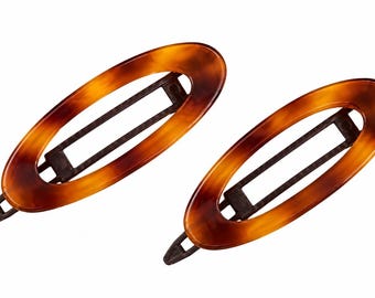 May include: Two oval-shaped hair clips featuring a glossy, mottled brown and amber tortoiseshell pattern. Each clip has a central open cutout design and a dark brown plastic snap-style mechanism visible underneath the decorative outer frame.