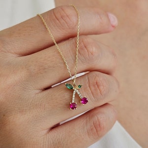 May include: A gold chain necklace with a pendant of two red cherries with green leaves and clear stones.