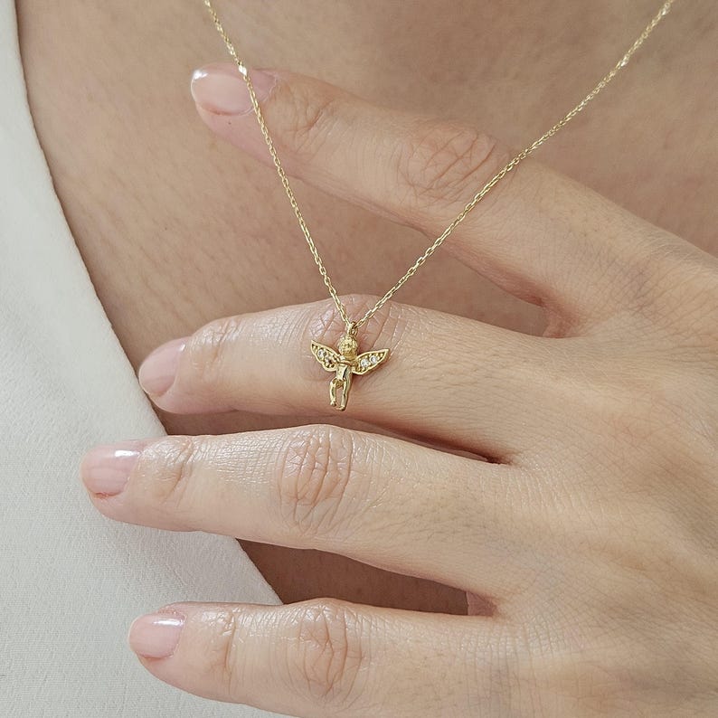May include: A gold chain necklace with a small angel pendant. The angel has wings and is adorned with tiny clear stones.
