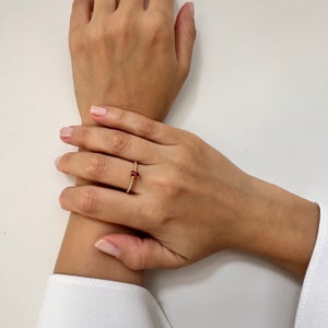 Op de afbeelding: Een gouden ring met een kleine rode edelsteen. De ring zit aan een vinger.
