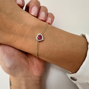 May include: A gold chain bracelet with a red heart-shaped pendant accented with clear cubic zirconia stones.