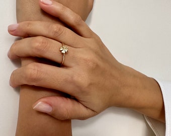 Puede incluir: Un anillo dorado con un diseño de trébol de cuatro hojas. El anillo tiene una banda delgada y se usa en un dedo. La imagen muestra un primer plano de una mano con el anillo, sobre un fondo blanco.