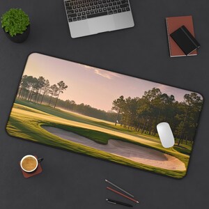 May include: A rectangular desk mat featuring a golf course scene with a sand trap, green grass, and tall trees under a soft, pink-hued sky. A white computer mouse rests on the mat, alongside a cup of coffee and a laptop.