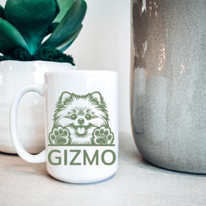 K&ouml;nnte beinhalten: Wei&szlig;e Keramik-Kaffeetasse mit einer gr&uuml;nen Illustration eines Zwergspitz-Hundes, der &uuml;ber den Rand schaut. Die Tasse tr&auml;gt den Schriftzug "Gizmo" in Gr&uuml;n unter dem Hund.