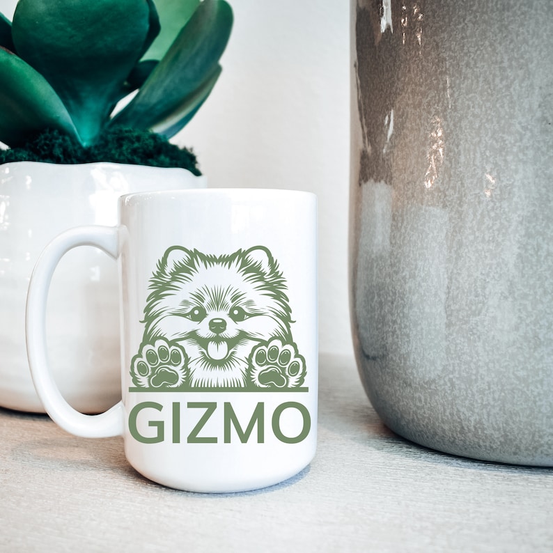 K&ouml;nnte beinhalten: Wei&szlig;e Keramik-Kaffeetasse mit einer gr&uuml;nen Illustration eines Zwergspitz-Hundes, der &uuml;ber den Rand schaut. Die Tasse tr&auml;gt den Schriftzug "Gizmo" in Gr&uuml;n unter dem Hund.